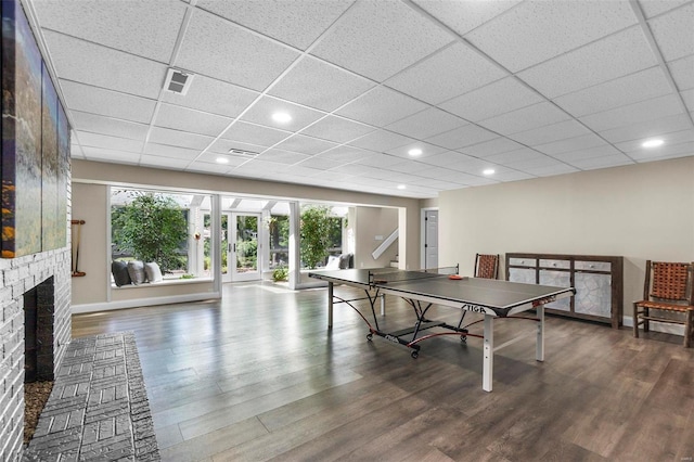 recreation room with a paneled ceiling, a brick fireplace, wood finished floors, and visible vents