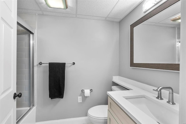 bathroom featuring toilet, enclosed tub / shower combo, vanity, a drop ceiling, and baseboards