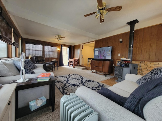 living room with a wood stove, ceiling fan, wooden walls, and light carpet