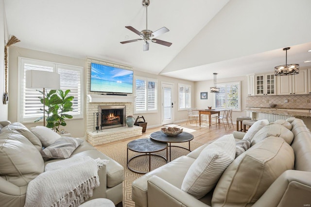 living room featuring a ceiling fan, baseboards, high vaulted ceiling, light wood-style floors, and a brick fireplace