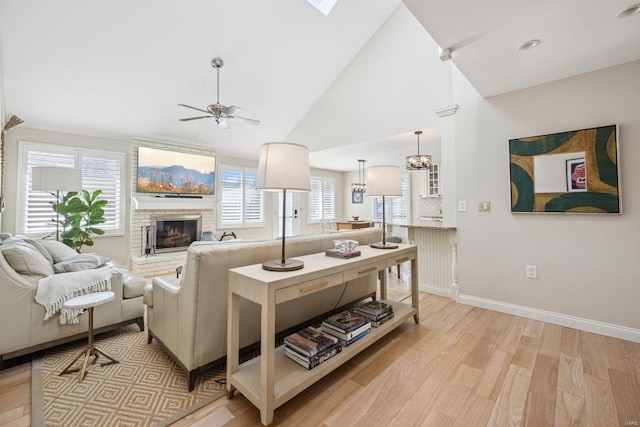 living area with baseboards, ceiling fan, light wood-type flooring, a fireplace, and high vaulted ceiling