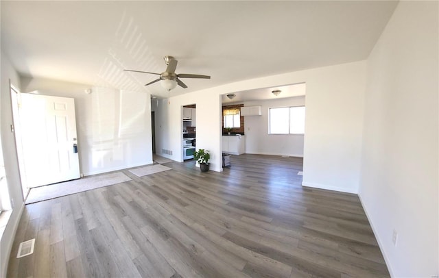 unfurnished living room with a wall unit AC, visible vents, baseboards, dark wood finished floors, and ceiling fan