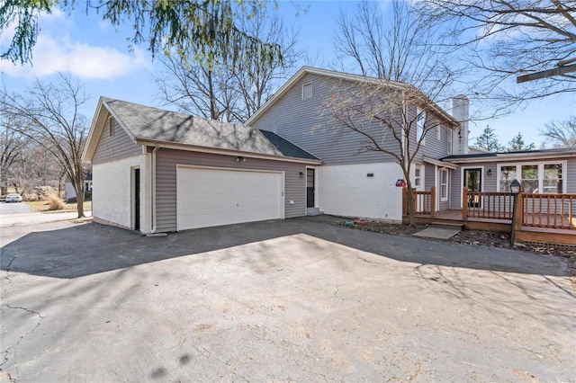 view of home's exterior with a deck, an attached garage, brick siding, driveway, and a chimney