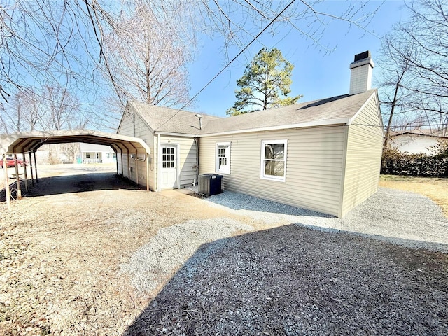 back of house with a shingled roof, a chimney, gravel driveway, central air condition unit, and a detached carport