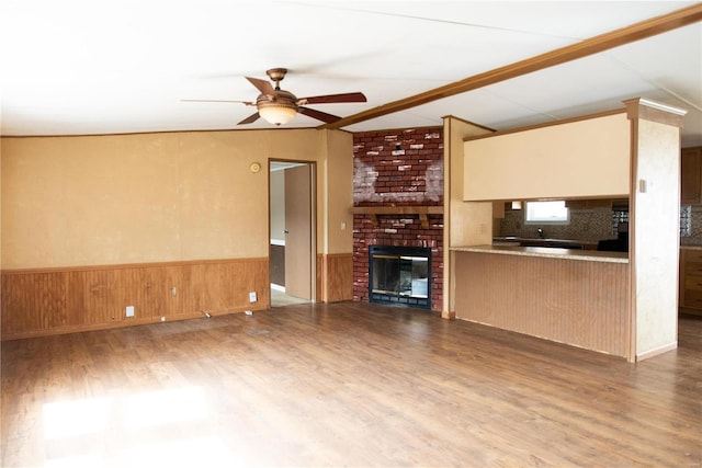 unfurnished living room featuring a wainscoted wall, a brick fireplace, a ceiling fan, and wood finished floors