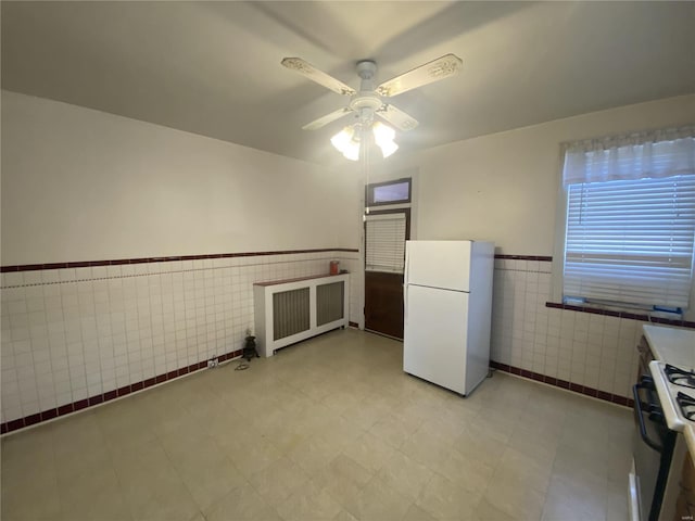 kitchen featuring a ceiling fan, gas range, a wainscoted wall, freestanding refrigerator, and tile walls