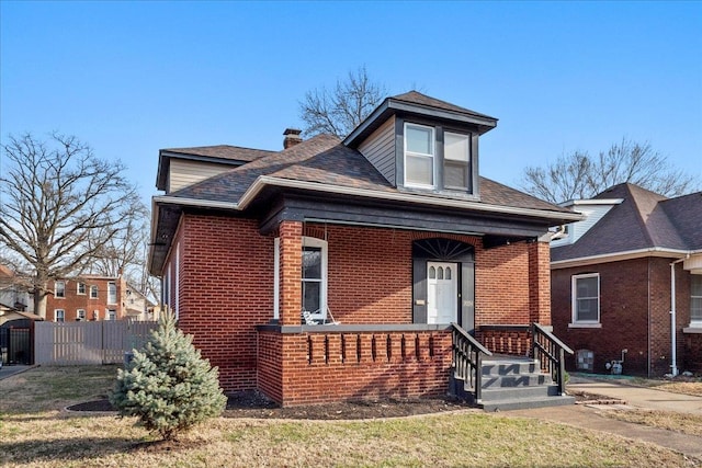 bungalow-style house with covered porch, brick siding, a shingled roof, fence, and a front lawn