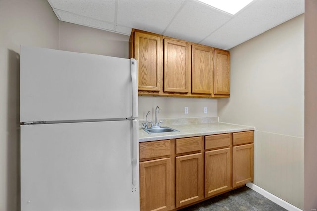 kitchen with a drop ceiling, brown cabinets, freestanding refrigerator, light countertops, and a sink