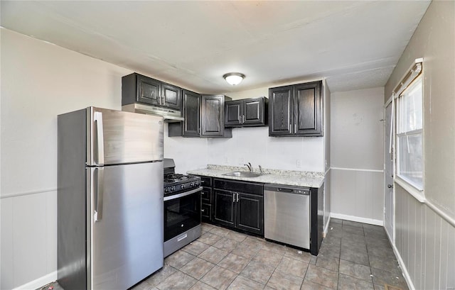 kitchen with a wainscoted wall, stainless steel appliances, dark cabinetry, under cabinet range hood, and a sink