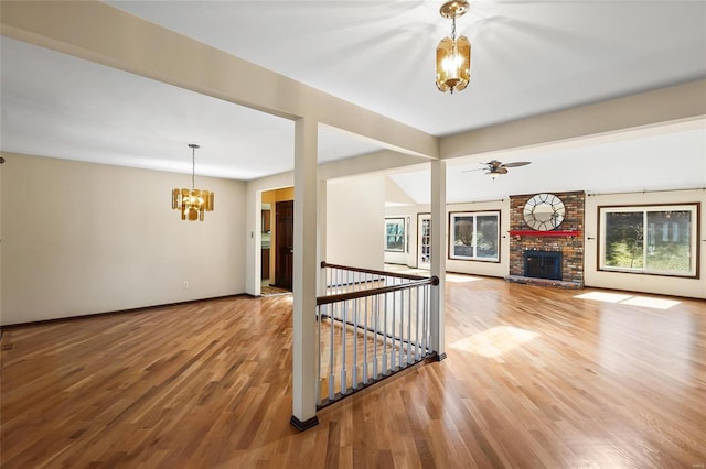 unfurnished living room with ceiling fan with notable chandelier, a brick fireplace, wood finished floors, and baseboards