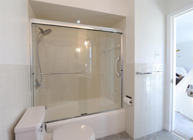 ensuite bathroom featuring toilet, bath / shower combo with glass door, tile walls, and wainscoting