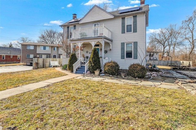 view of front facade featuring a chimney, a porch, a front yard, roof mounted solar panels, and a balcony