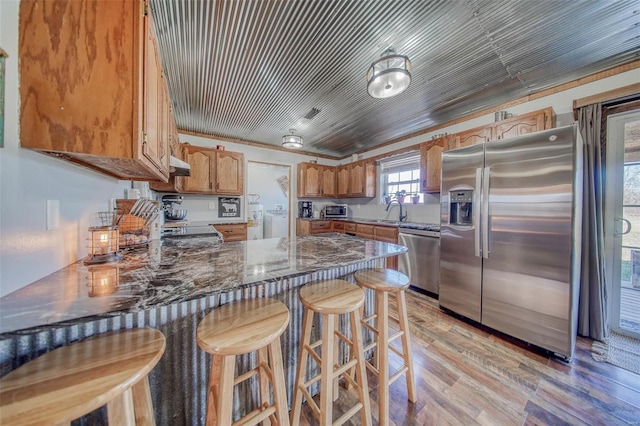 kitchen featuring brown cabinetry, a peninsula, stainless steel appliances, under cabinet range hood, and light wood-type flooring