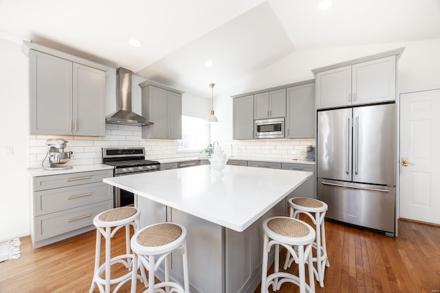 kitchen featuring lofted ceiling, gray cabinets, appliances with stainless steel finishes, wall chimney range hood, and a kitchen bar