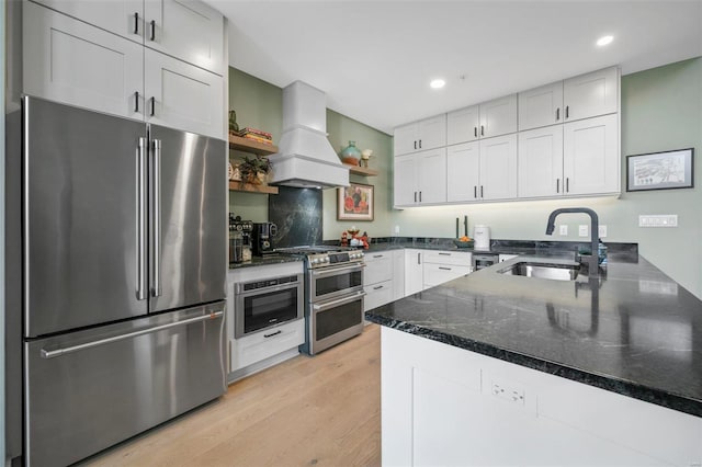 kitchen featuring stainless steel appliances, premium range hood, a sink, dark stone counters, and light wood finished floors