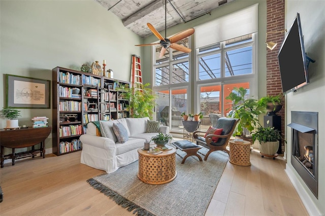 living area with a towering ceiling, a warm lit fireplace, ceiling fan, and wood finished floors