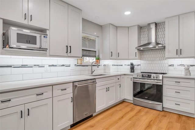 kitchen with a sink, wall chimney range hood, light wood-style floors, appliances with stainless steel finishes, and light countertops