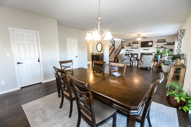 dining space with baseboards, stairway, wood finished floors, a fireplace, and ceiling fan with notable chandelier