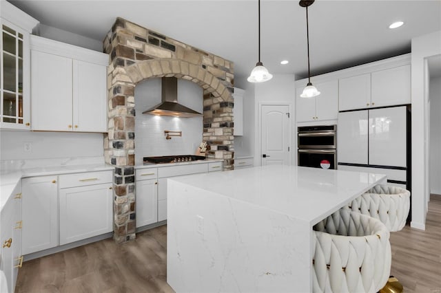 kitchen with stainless steel appliances, light wood finished floors, glass insert cabinets, and wall chimney range hood