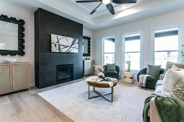 living area featuring light wood-style flooring, a fireplace, a tray ceiling, and a ceiling fan