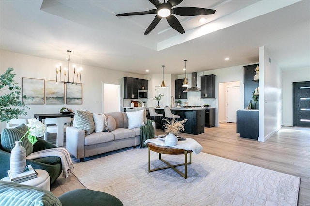 living area featuring recessed lighting, ceiling fan with notable chandelier, baseboards, light wood-style floors, and a raised ceiling