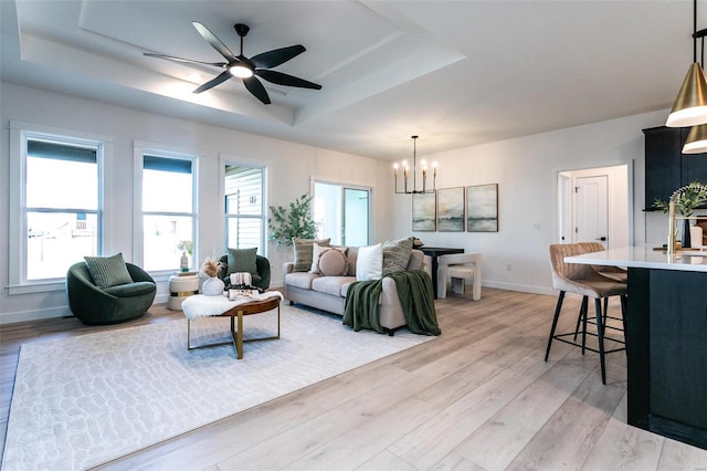 living area with light wood-style flooring, a tray ceiling, baseboards, and ceiling fan with notable chandelier