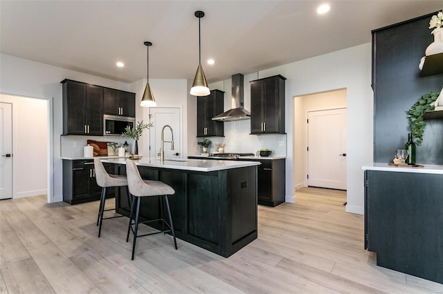 kitchen featuring a center island with sink, decorative backsplash, wall chimney exhaust hood, light countertops, and a sink