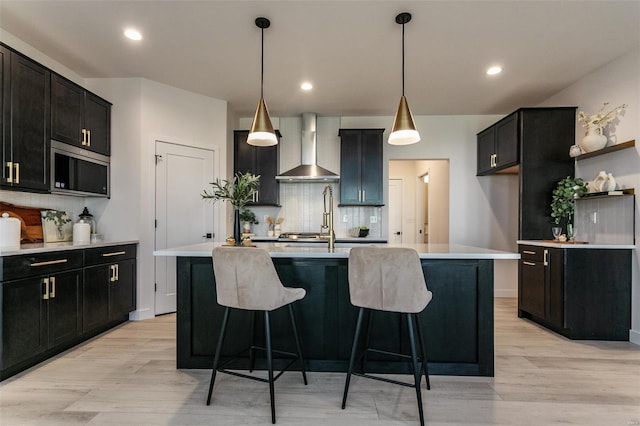 kitchen with light countertops, wall chimney range hood, dark cabinetry, stainless steel microwave, and a center island with sink