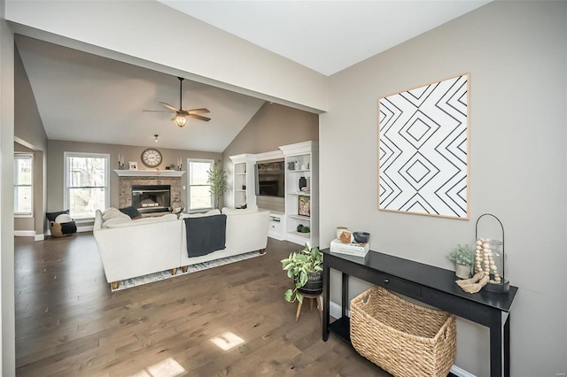 living area featuring dark wood finished floors, baseboards, vaulted ceiling, a ceiling fan, and a glass covered fireplace