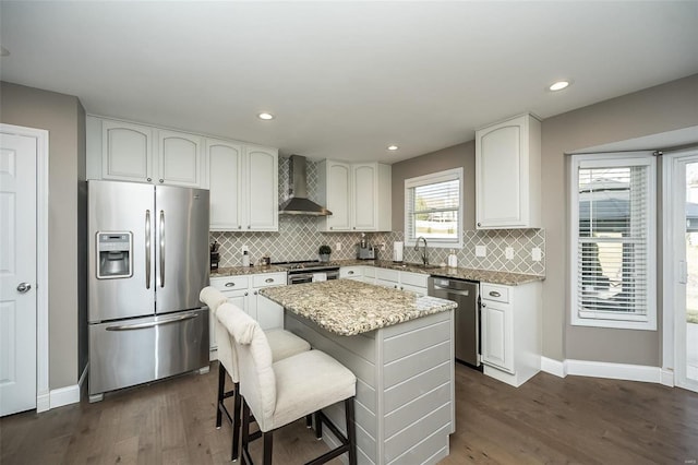 kitchen with dark wood-style flooring, appliances with stainless steel finishes, a sink, wall chimney range hood, and light stone countertops