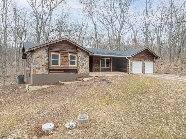 view of front facade with driveway, an attached garage, a shingled roof, stone siding, and central air condition unit