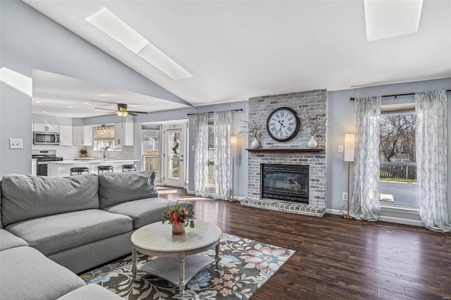 living area featuring wood finished floors, baseboards, a fireplace, ceiling fan, and lofted ceiling with skylight