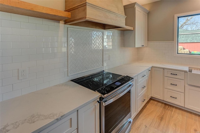 kitchen with light stone counters, premium range hood, light wood-style floors, stainless steel range with electric cooktop, and backsplash