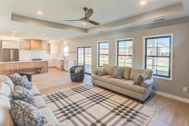 living room with light wood-style floors, recessed lighting, a raised ceiling, and baseboards