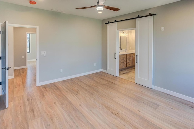 unfurnished bedroom with a barn door, a ceiling fan, ensuite bath, light wood-type flooring, and baseboards