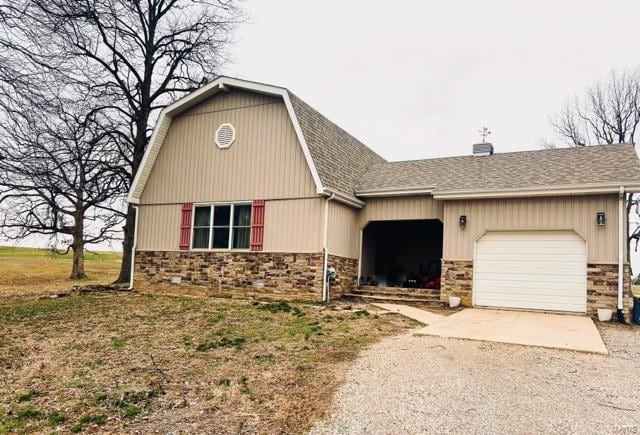 view of front facade featuring a shingled roof, a gambrel roof, concrete driveway, a garage, and stone siding