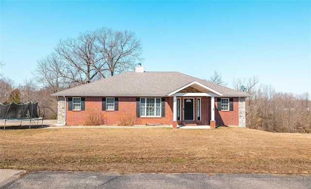 ranch-style house featuring a shingled roof, a chimney, a front lawn, a trampoline, and brick siding