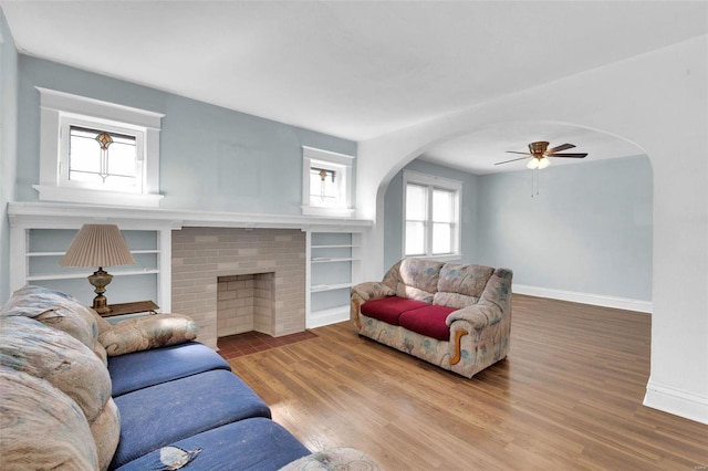 living room featuring a ceiling fan, wood finished floors, arched walkways, baseboards, and a brick fireplace