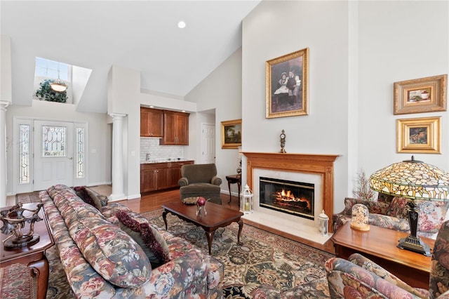 living area with wood finished floors, baseboards, ornate columns, high vaulted ceiling, and a glass covered fireplace