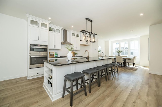 kitchen featuring dark countertops, wall chimney range hood, a breakfast bar, an island with sink, and light wood-style floors