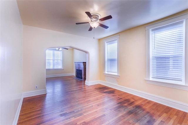 unfurnished living room with a ceiling fan, wood finished floors, arched walkways, baseboards, and a brick fireplace