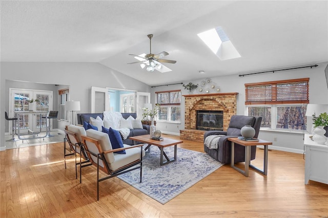 living area featuring a ceiling fan, baseboards, vaulted ceiling with skylight, light wood-style floors, and a brick fireplace