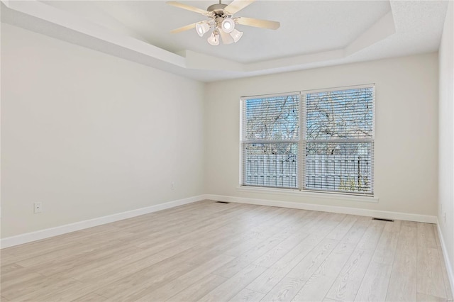 empty room featuring a raised ceiling, visible vents, light wood-style floors, a ceiling fan, and baseboards