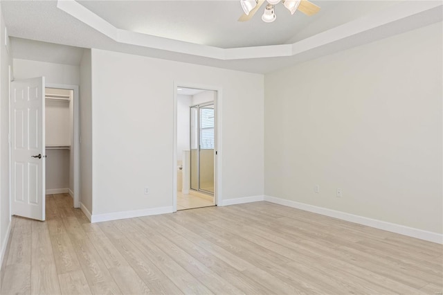 unfurnished bedroom featuring light wood finished floors, baseboards, ensuite bathroom, a tray ceiling, and a walk in closet