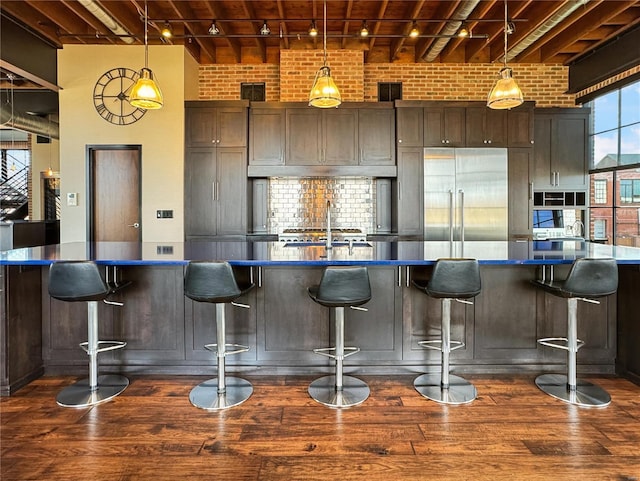 kitchen featuring dark countertops, brick wall, stainless steel built in refrigerator, dark wood-style floors, and a sink