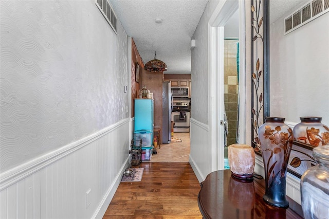 hall featuring a wainscoted wall, wood finished floors, visible vents, and a textured ceiling