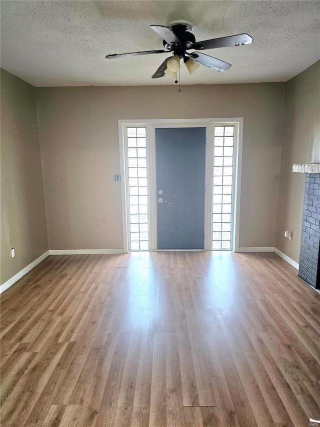 unfurnished room with a textured ceiling, wood finished floors, baseboards, a brick fireplace, and ceiling fan