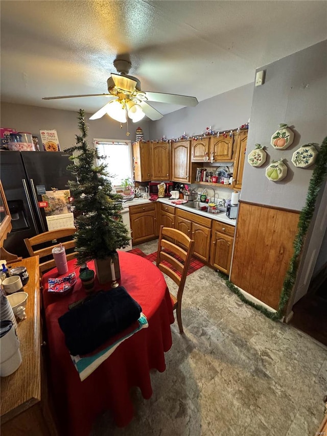 kitchen with wood walls, brown cabinets, a ceiling fan, a sink, and wainscoting