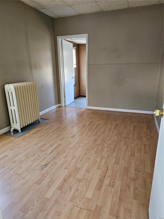 empty room with light wood-style flooring, radiator heating unit, baseboards, and a drop ceiling