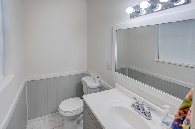 half bath with wainscoting, marble finish floor, toilet, and vanity